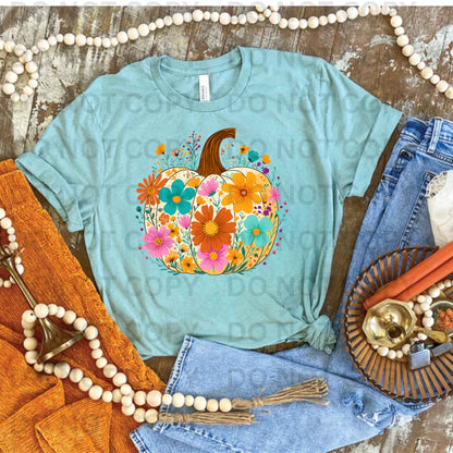 Light blue t-shirt with a colorful pumpkin design on a rustic wooden surface with decorative items.