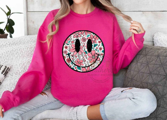 Person wearing a bright pink sweatshirt with a floral smiley face design, sitting on a couch.