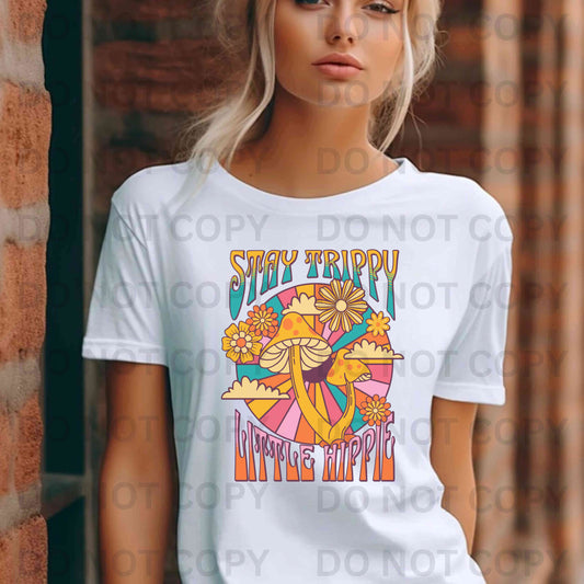 Person wearing a white t-shirt with a colorful graphic and text 'Stay Trippy Little Hippie' against a brick wall.